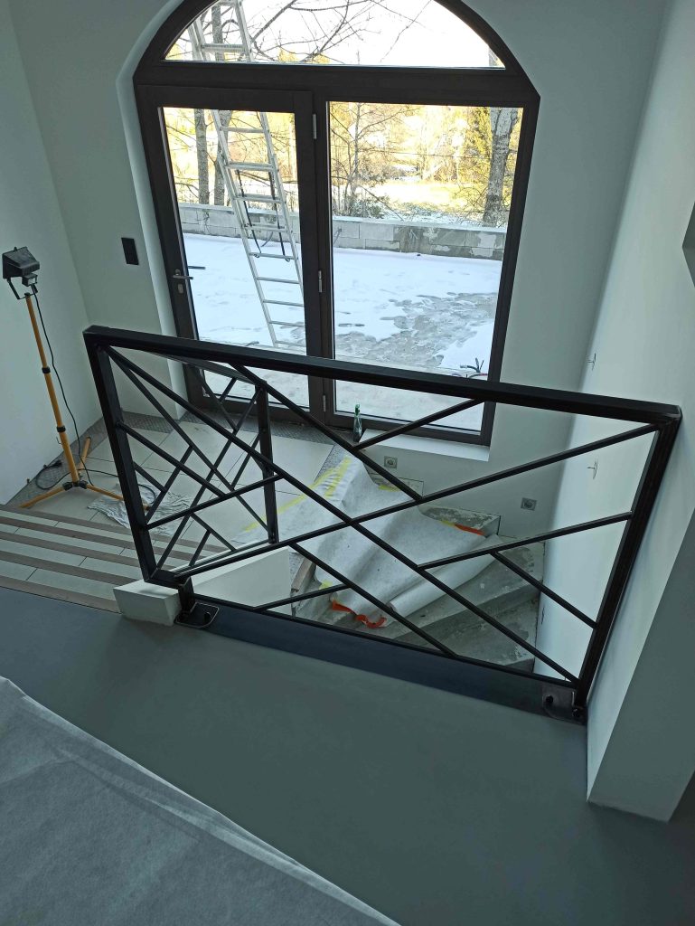Garde-corps intérieur à lignes croisées au bord du palier, devant une grande fenêtre cintrée.